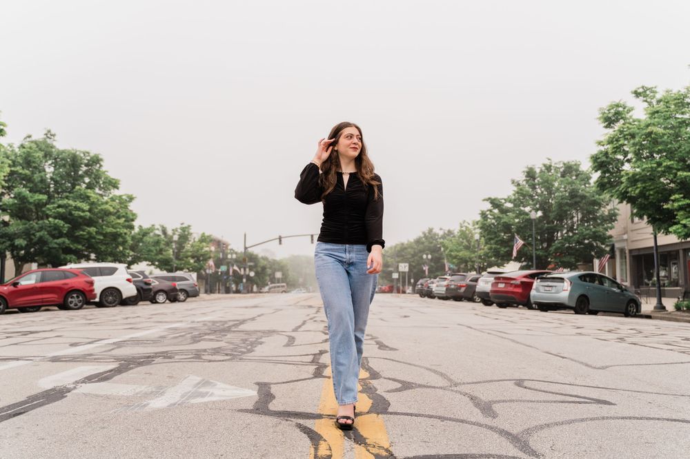 Walking shot in a parking lot wearing casual black top and jeans with cars in background.