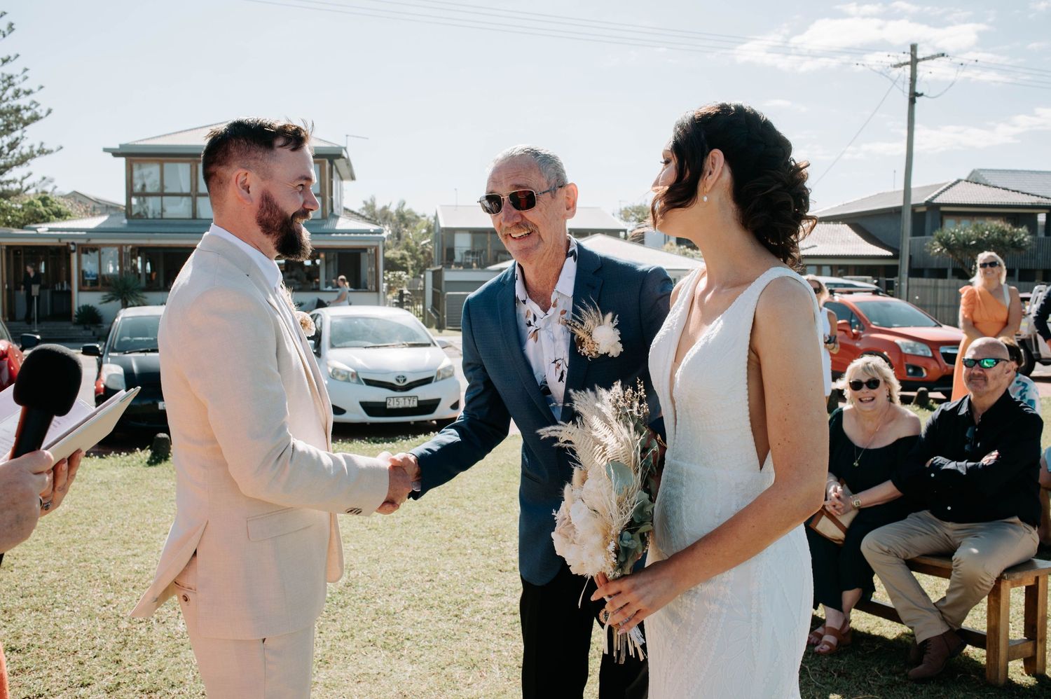 Outdoor wedding exchange under wooden arch with seated guests on lawn.