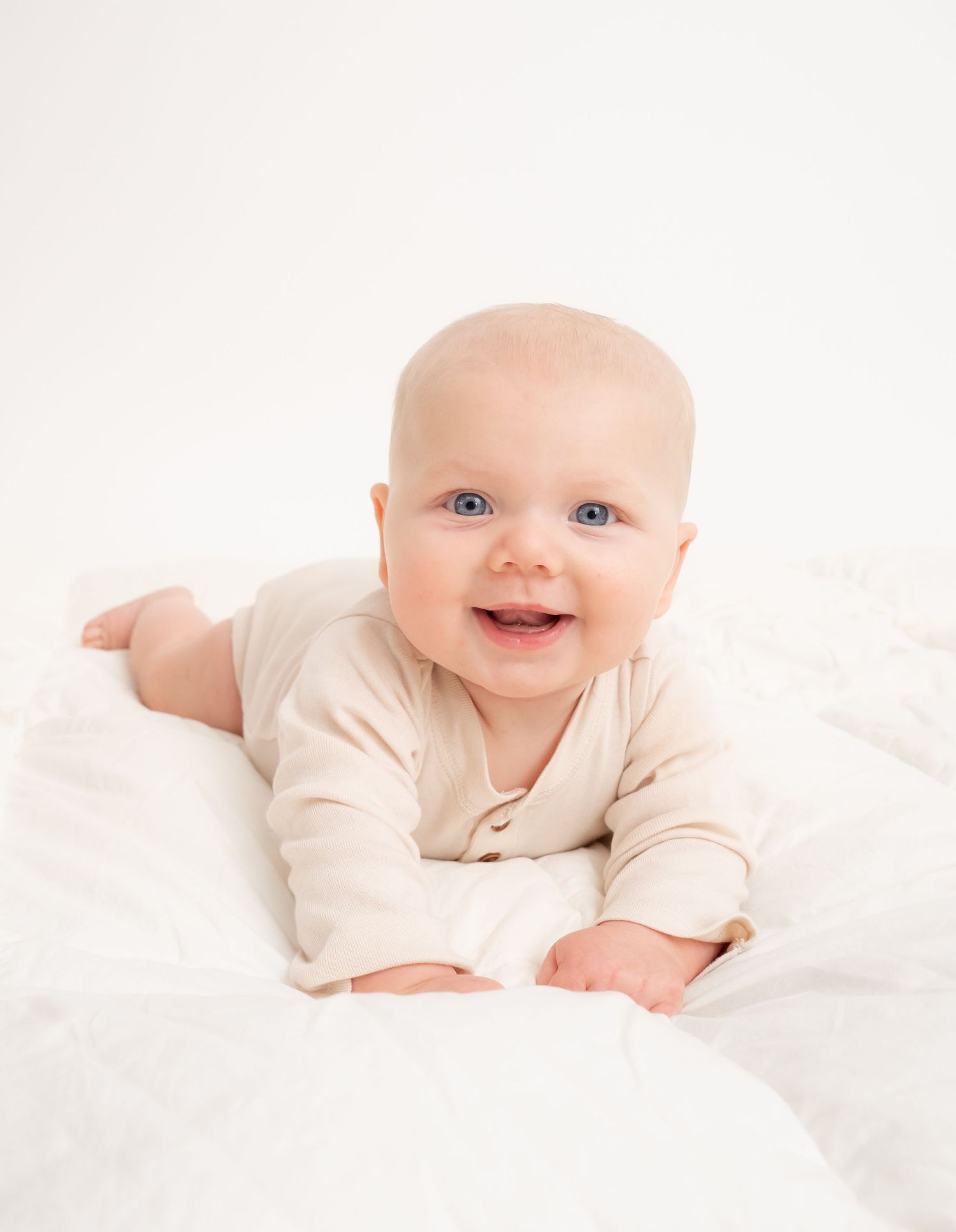 Smiling baby in cream colored onesie lying on white bedding.