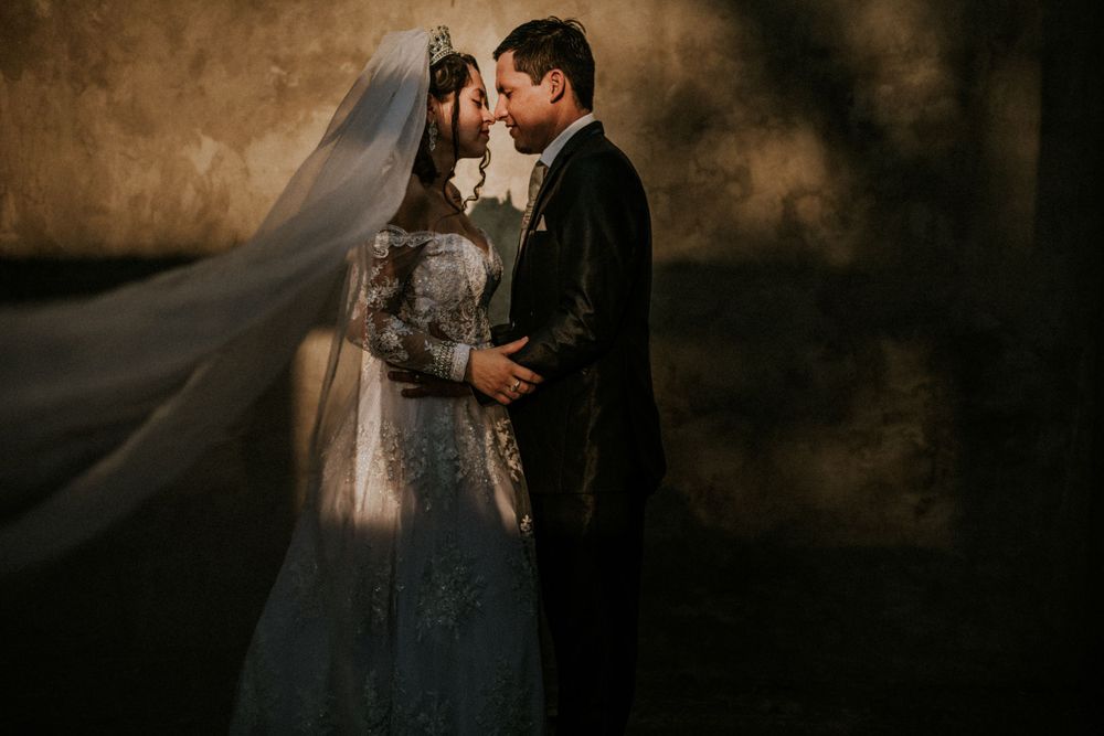 A couple embraces in a moody wedding portrait as the bride's long veil flows dramatically in dark atmospheric lighting.