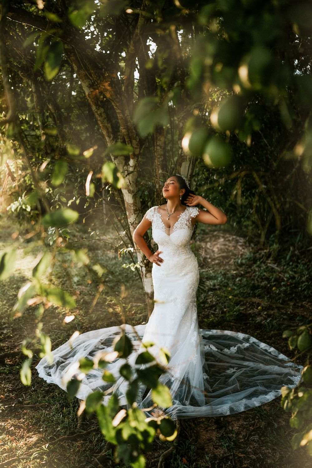 Sun-dappled wedding portrait showing a figure in an elegant lace gown with long train standing among garden foliage.