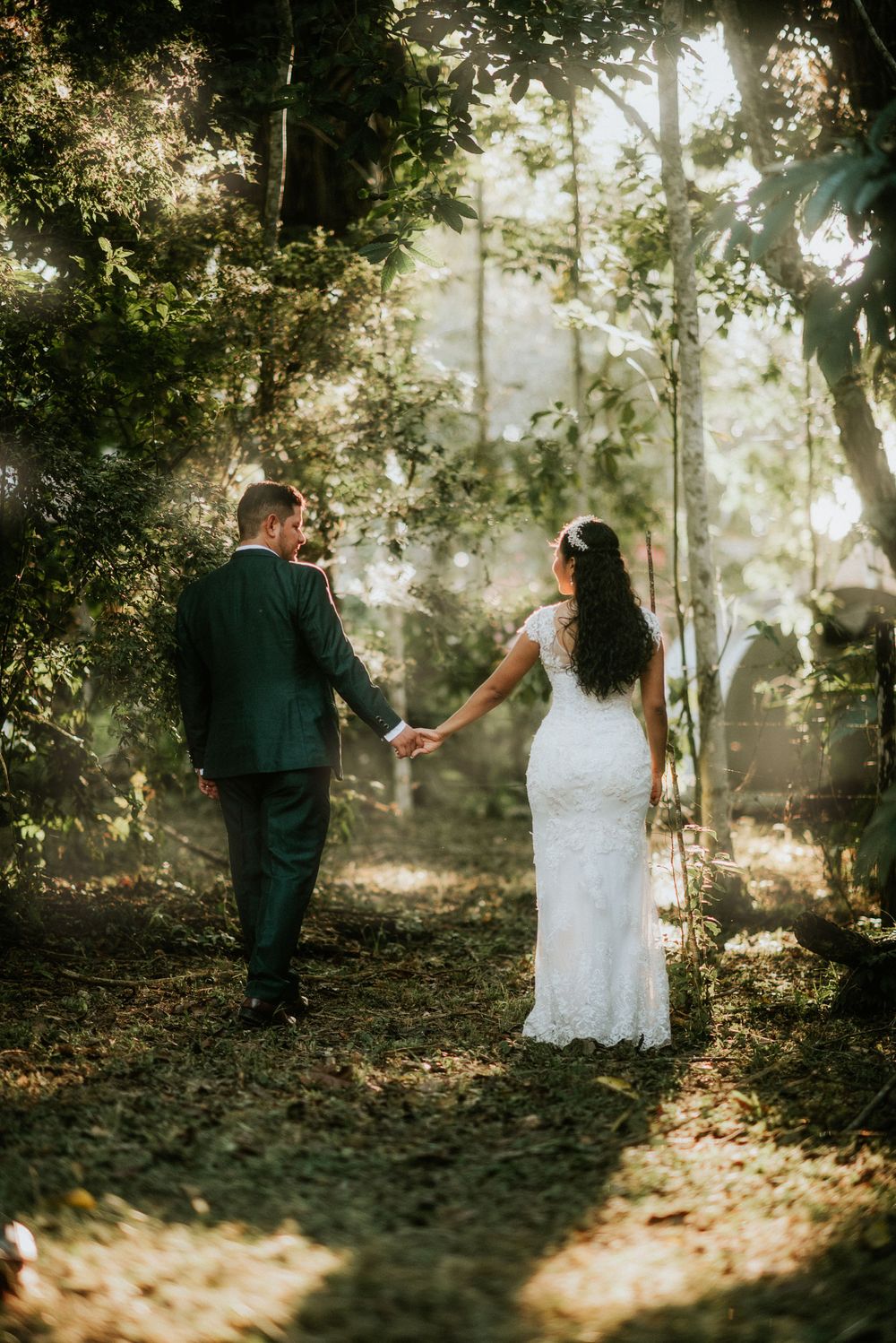 A couple in wedding attire holding hands while walking through a sunlit forest path surrounded by trees.