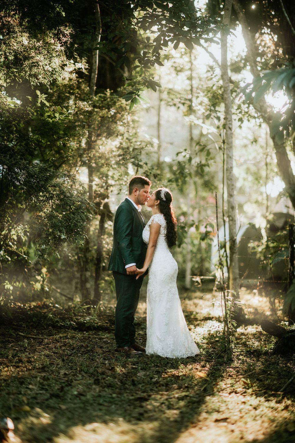 A couple in wedding attire embrace in a sunlit forest setting with rays of light filtering through the trees.