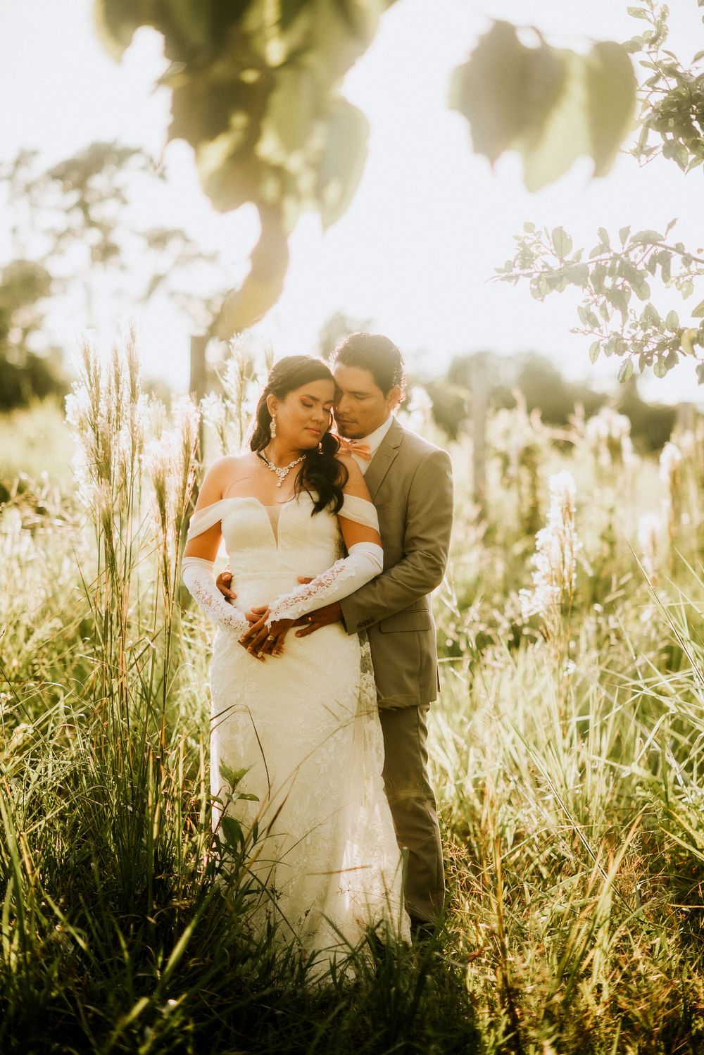Golden sunlit wedding photo of a couple embracing in tall meadow grass during sunset.