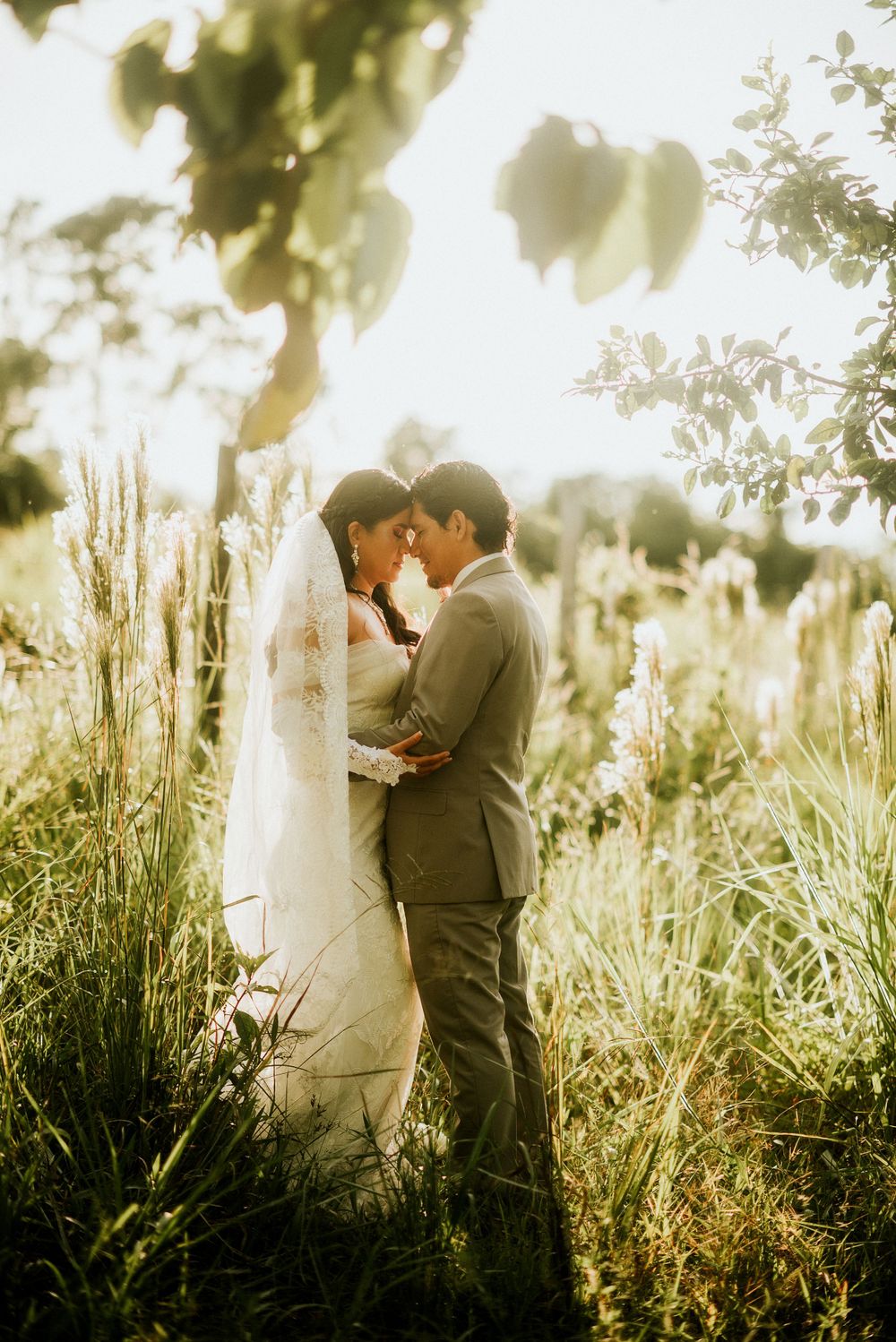 A romantic wedding photo taken in a sun-drenched field of tall grass and wildflowers at golden hour.