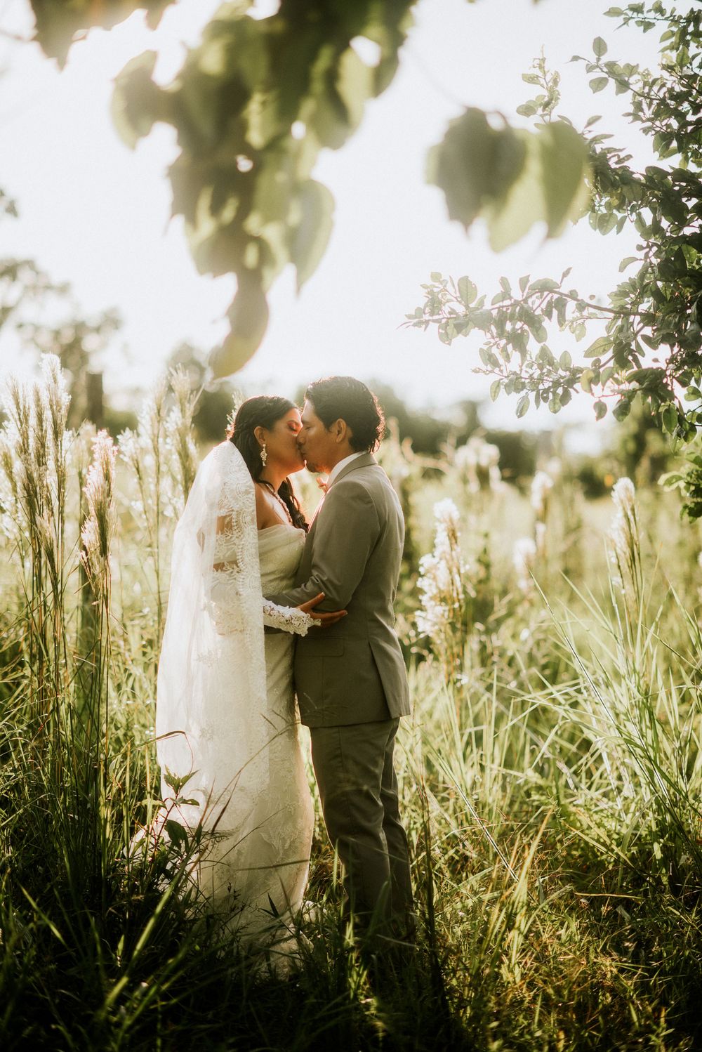 A romantic golden hour wedding portrait with a couple embracing in tall grass and sunlit trees.