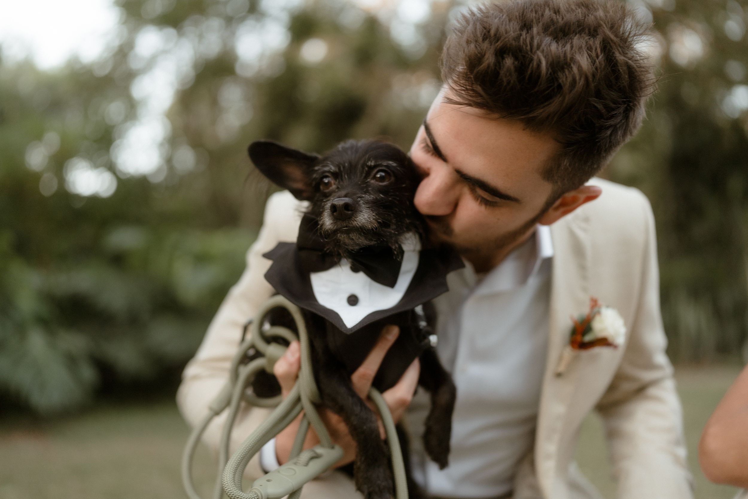 A groom in beige suit holds a small black dog wearing a bowtie tuxedo.