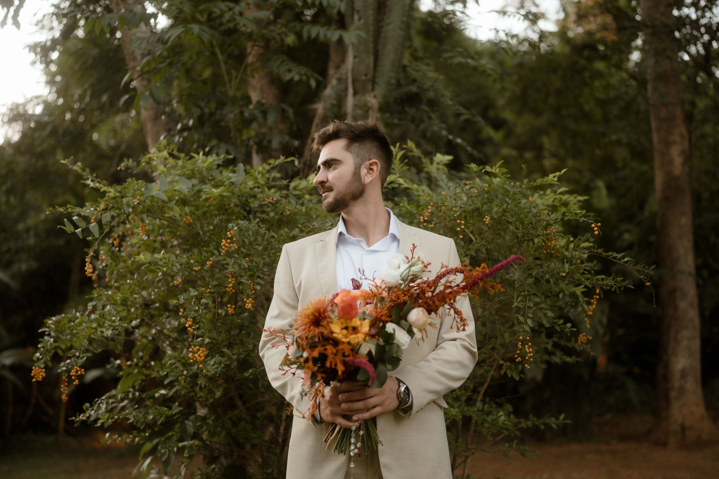 A person in a cream suit holds a stunning fall-colored bouquet against a lush garden backdrop.