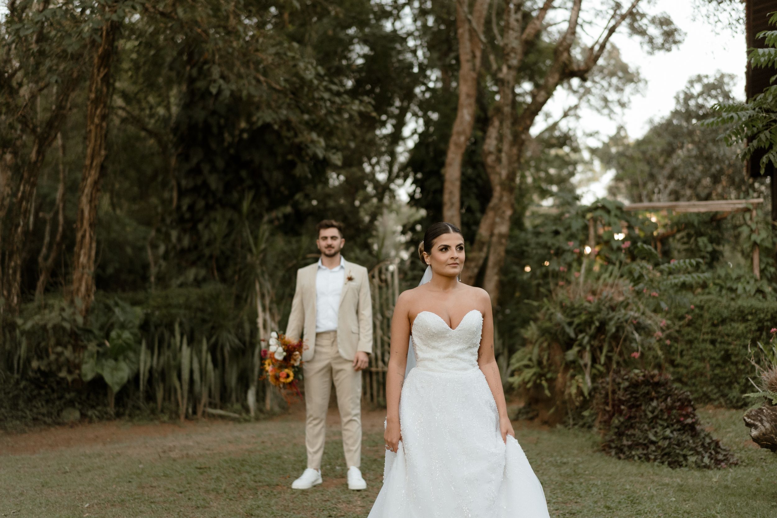 A wedding couple poses in a lush garden setting with greenery and string lights overhead.