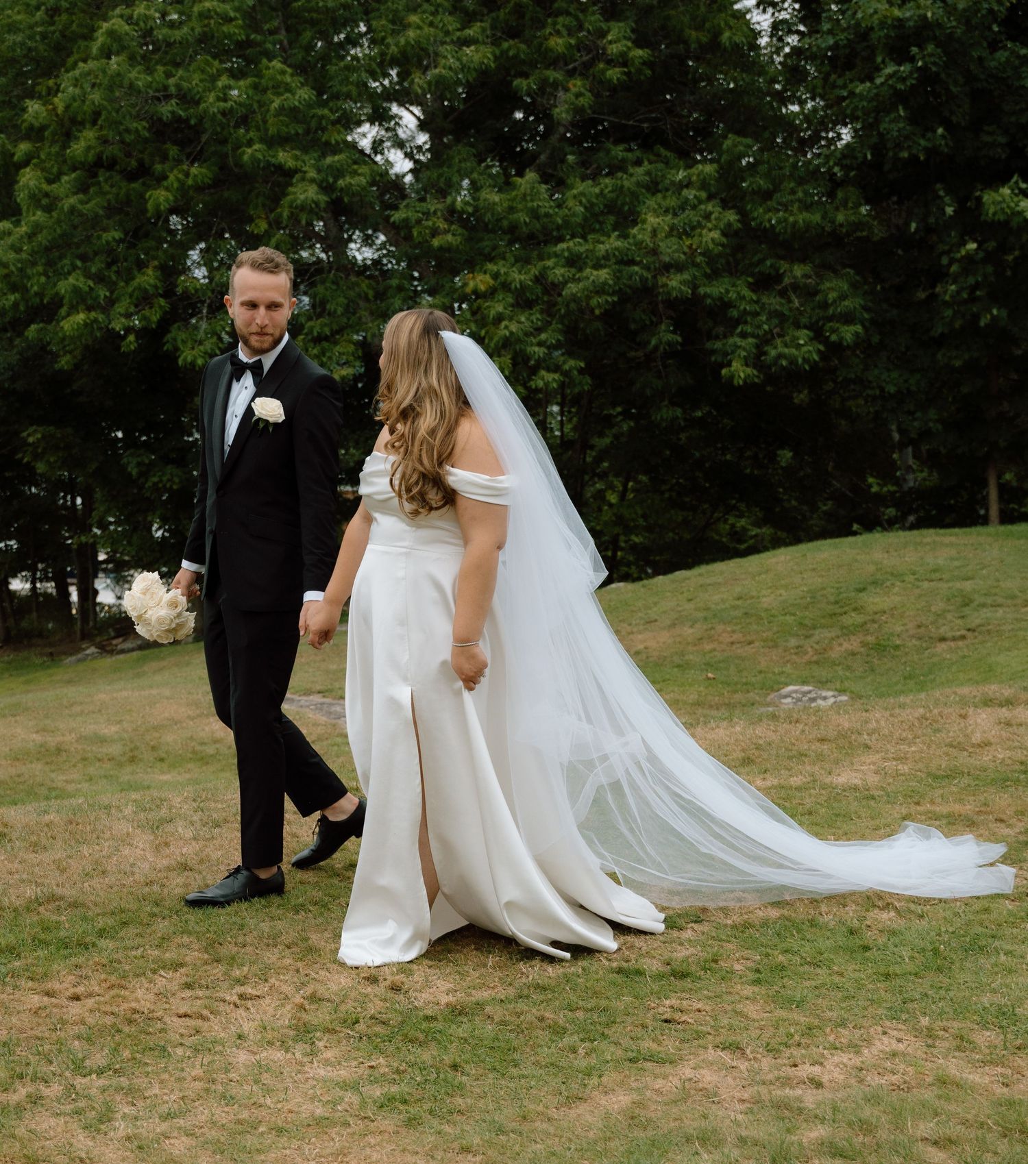 A wedding couple walks across a grassy hillside with flowing white dress and veil.