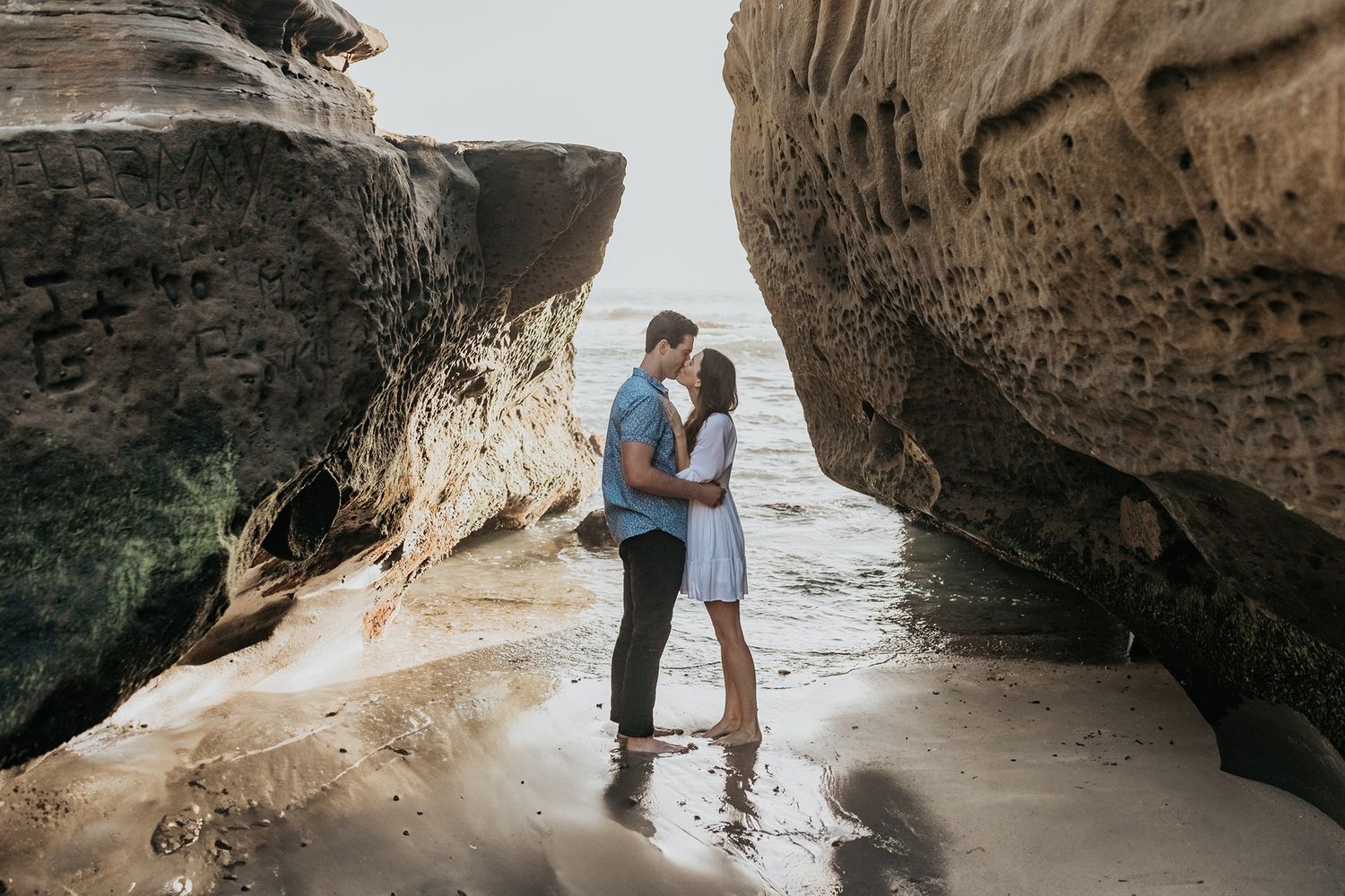 A couple embraces between large rock formations on a sandy beach during sunset at Windansea Beach in La Jolla.
