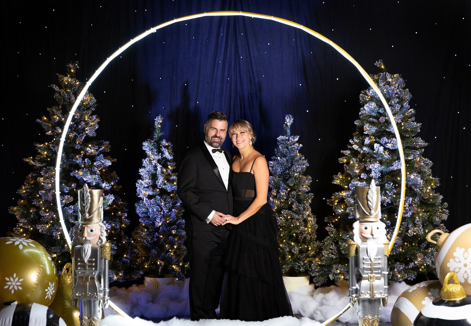 A couple poses under a white archway surrounded by decorated Christmas trees and ornaments at a formal winter event.