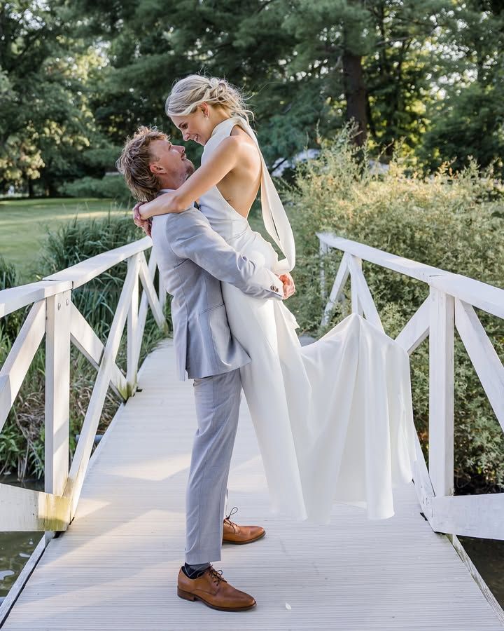 Romantic wedding couple embraces on white wooden bridge in lush garden setting during outdoor photoshoot.