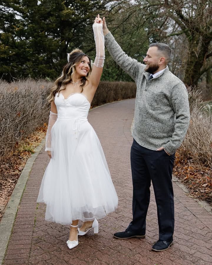 A couple dances playfully on a brick path surrounded by winter foliage, with one partner wearing a white dress.