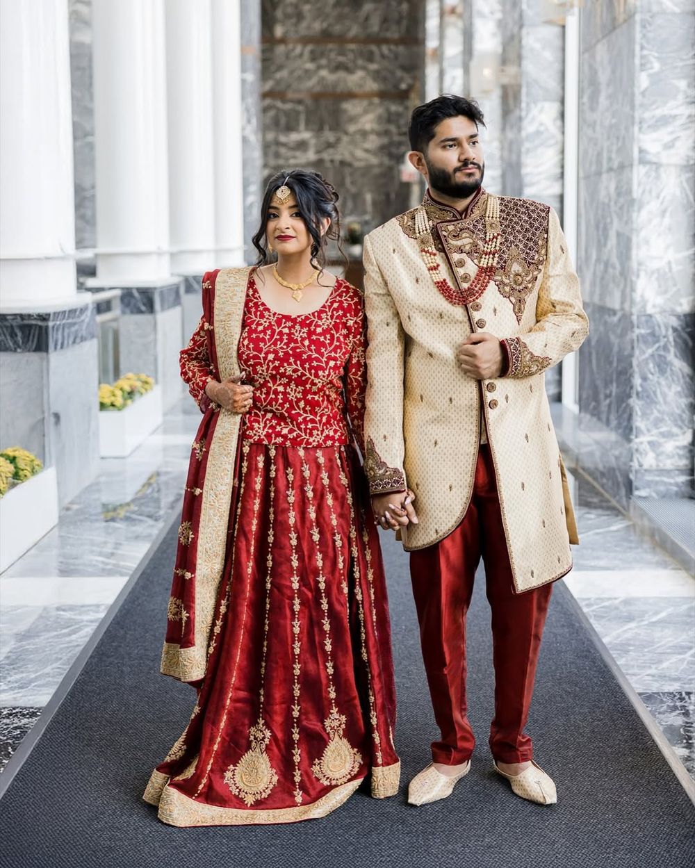 A couple in traditional South Asian wedding attire, with red and cream colored outfits, walking together in an elegant hallway.