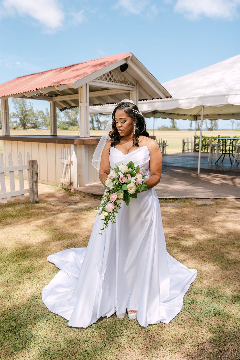 A bride in a flowing white dress poses with a bouquet in an outdoor garden setting with rustic structures.