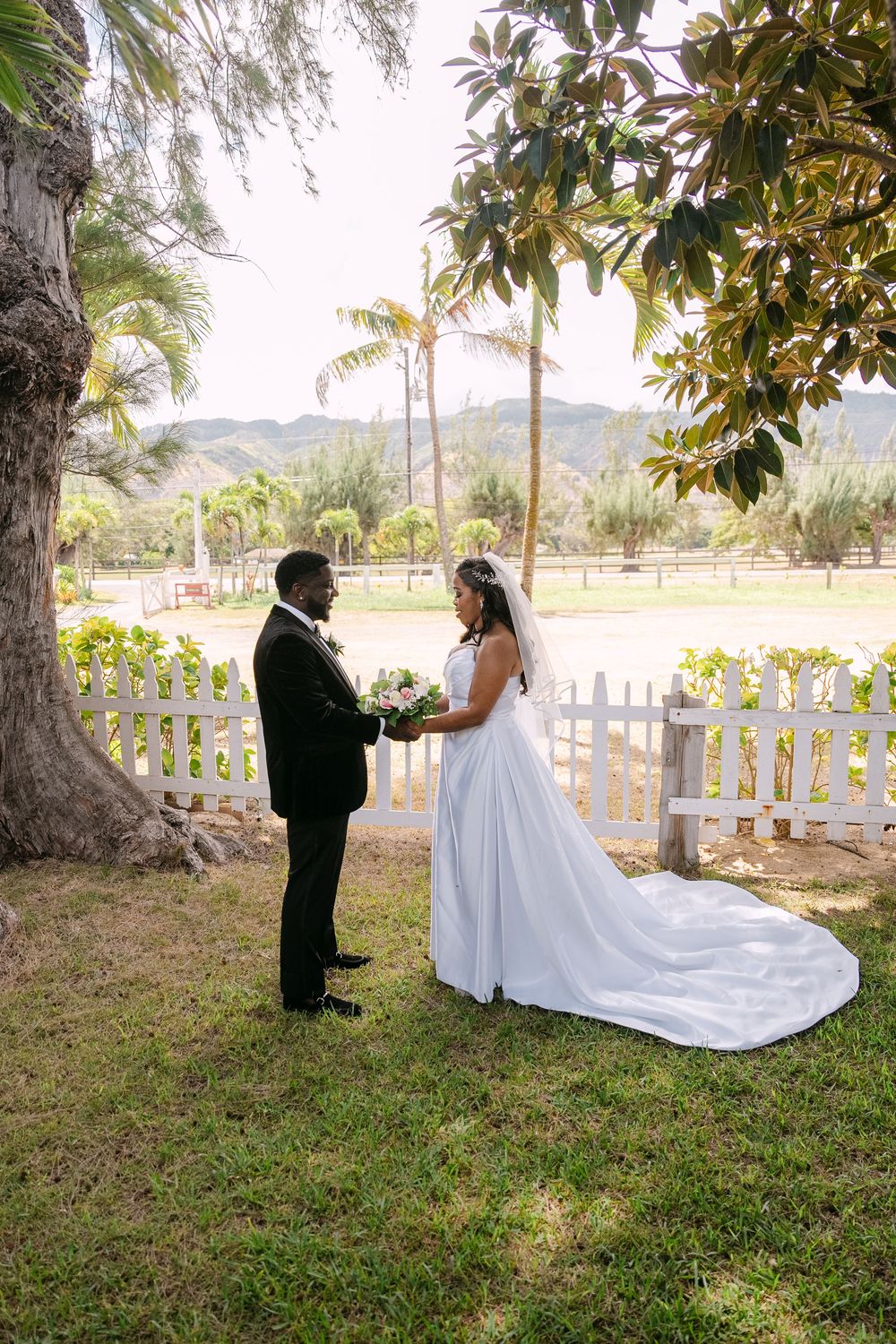 A wedding couple poses on a grassy lawn with white picket fences and palm trees in the background.