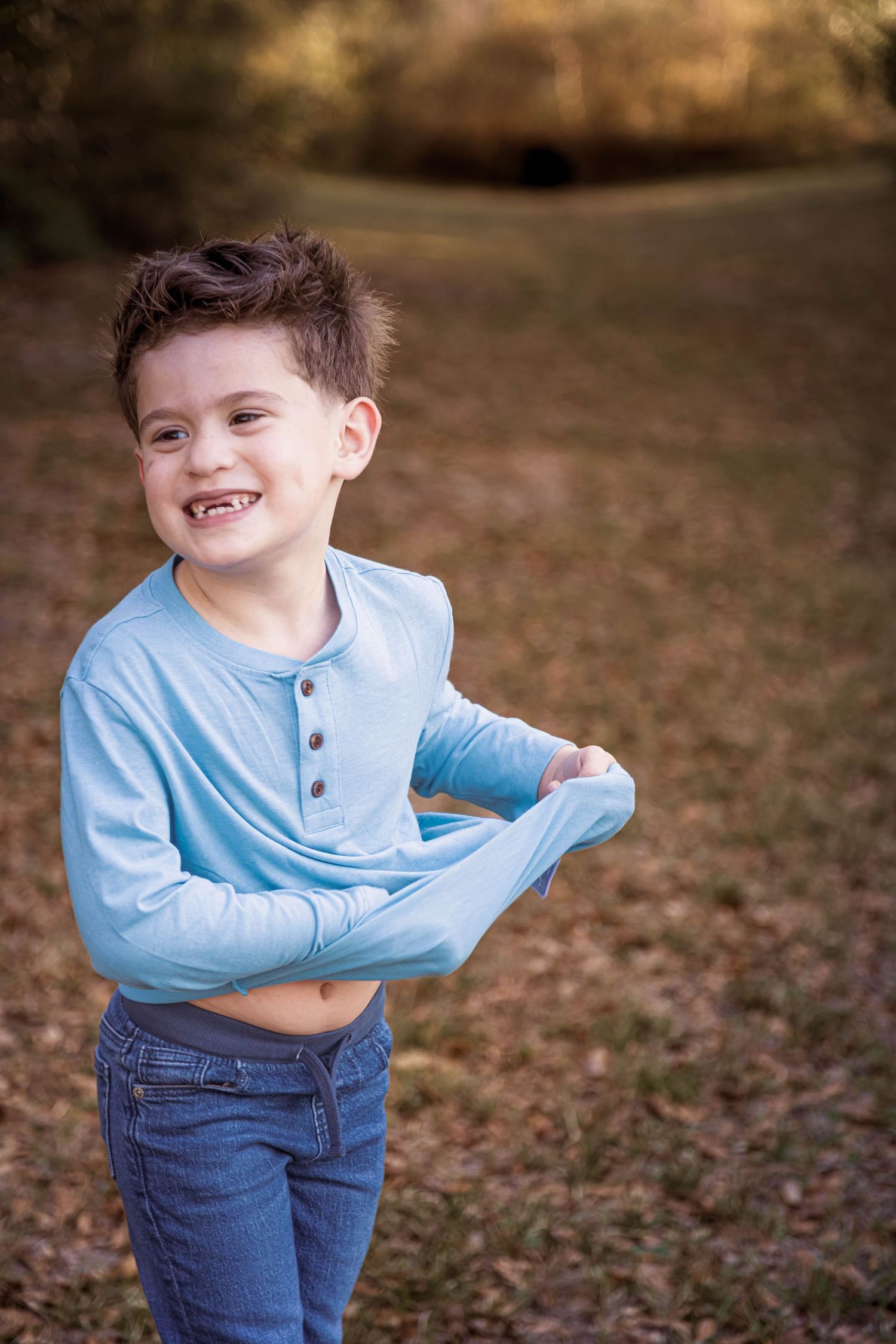A young child wearing a light blue henley shirt and jeans poses playfully outdoors on autumn leaves.