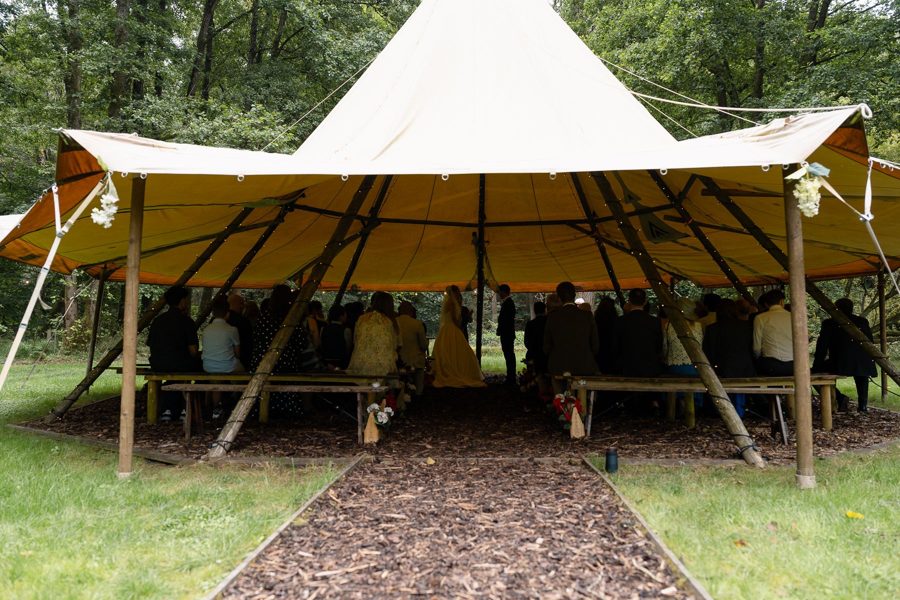 White outdoor event tent set up in a wooded area with mulch ground cover and green trees in the background.