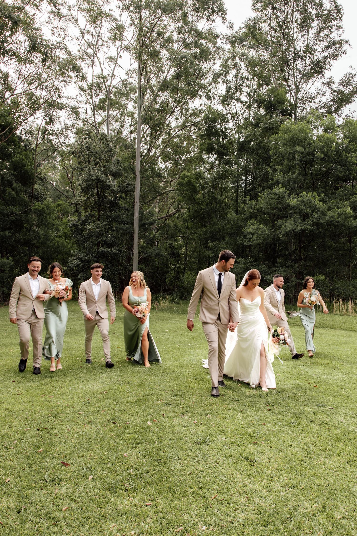Wedding party members playfully walk and pose across a grassy field surrounded by trees.