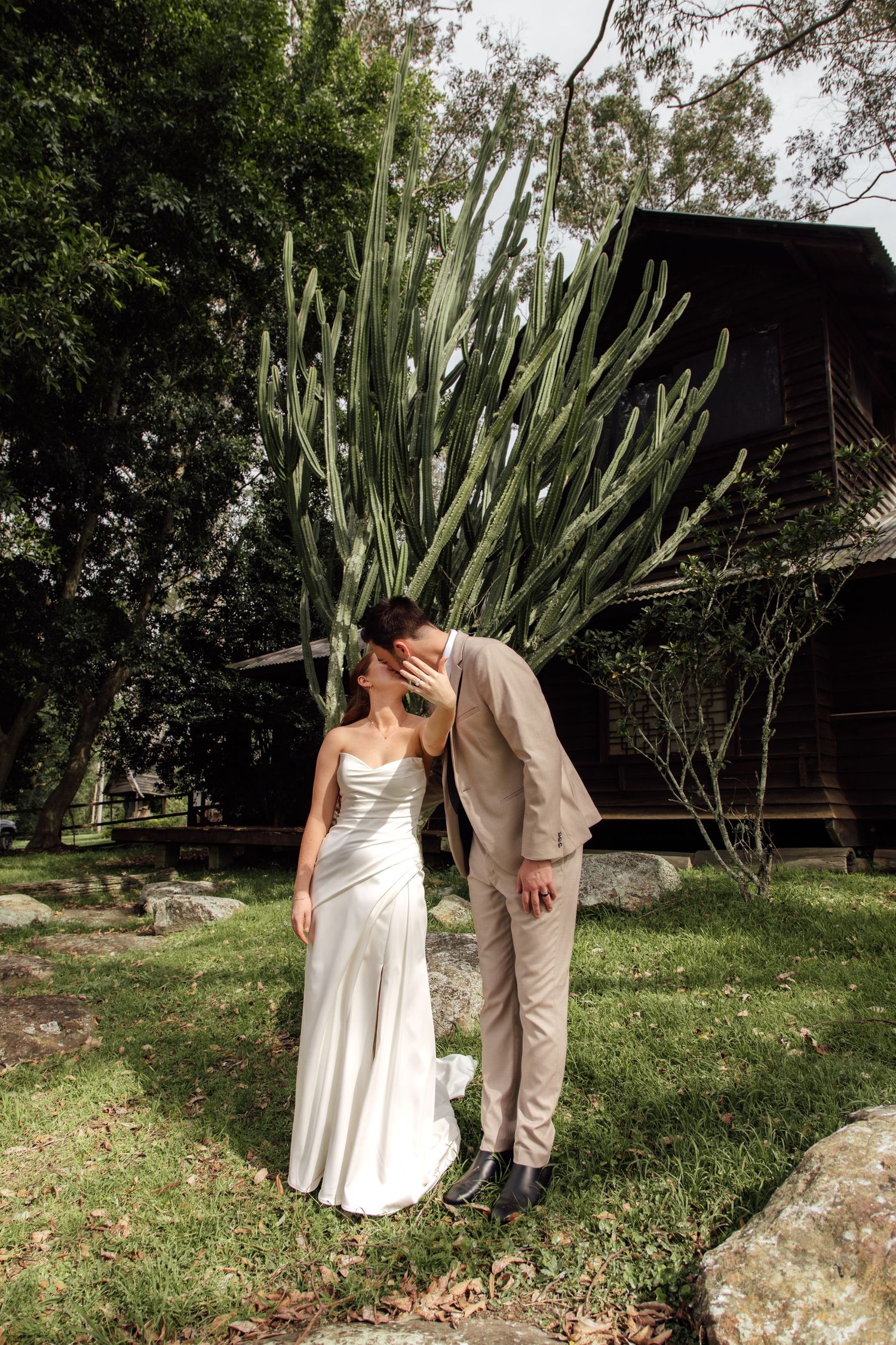 A couple in wedding attire shares intimate moments in a garden setting with tall cacti and trees in the background.