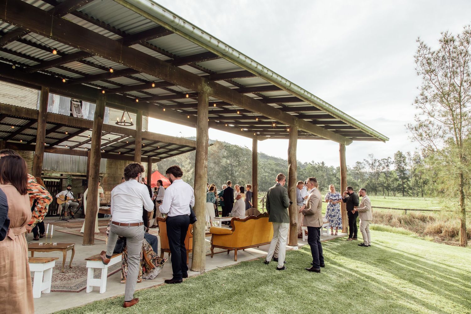 Wedding guests gather under a rustic covered pavilion during an outdoor ceremony in a scenic rural setting.