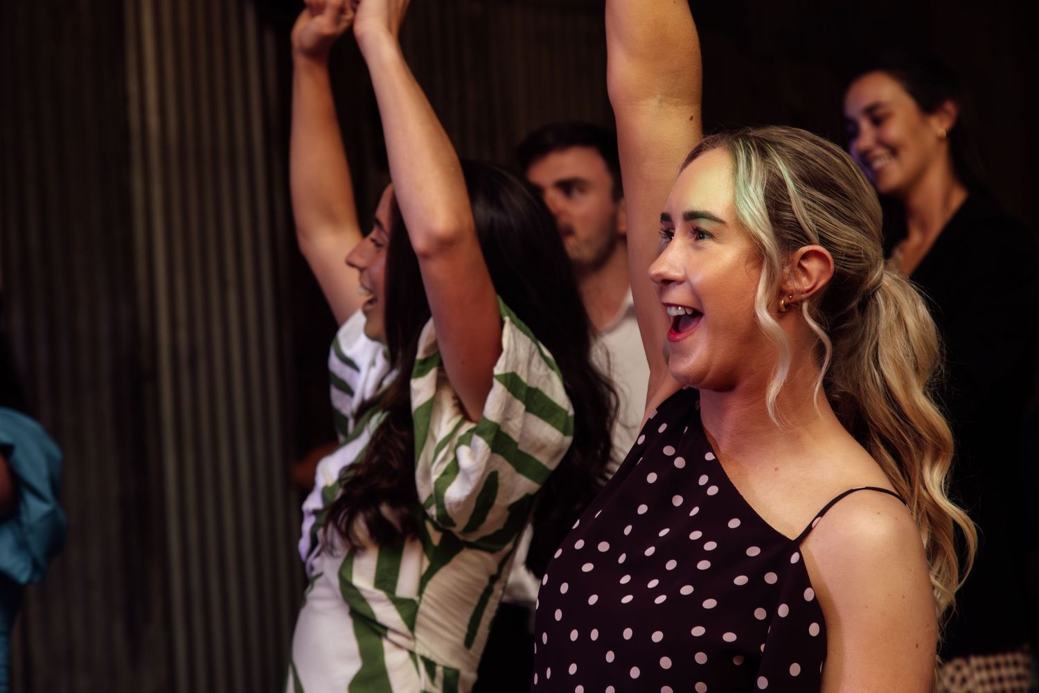 Wedding party members dancing enthusiastically at a reception with polka dot dresses visible.