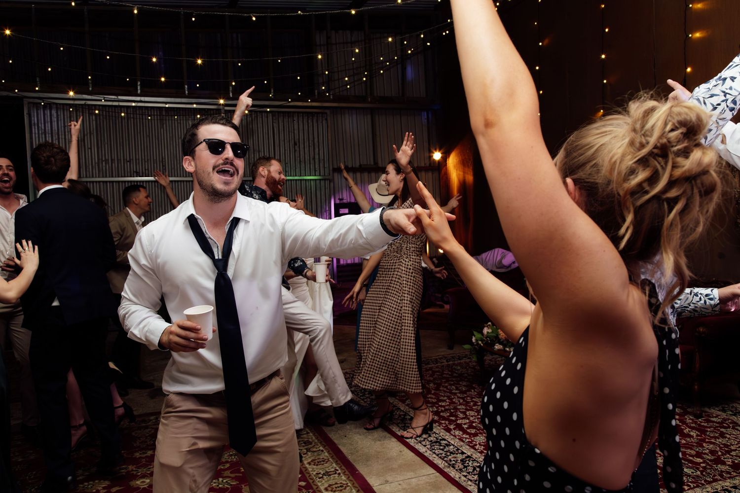 Wedding guests dance and celebrate on a dimly lit dance floor with string lights overhead during a reception party.