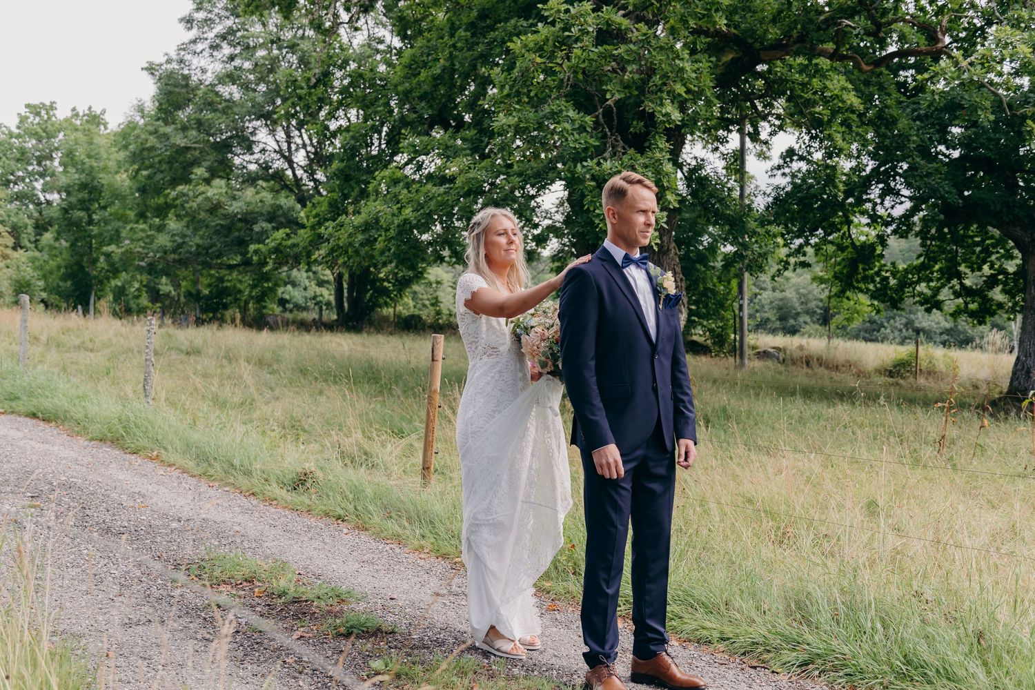 A couple stands together on a gravel path surrounded by lush green grass and trees during a summer wedding celebration.