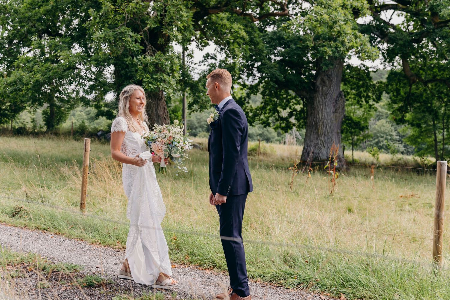 A couple in wedding attire stands together on a grassy hillside path during a sunny outdoor ceremony.