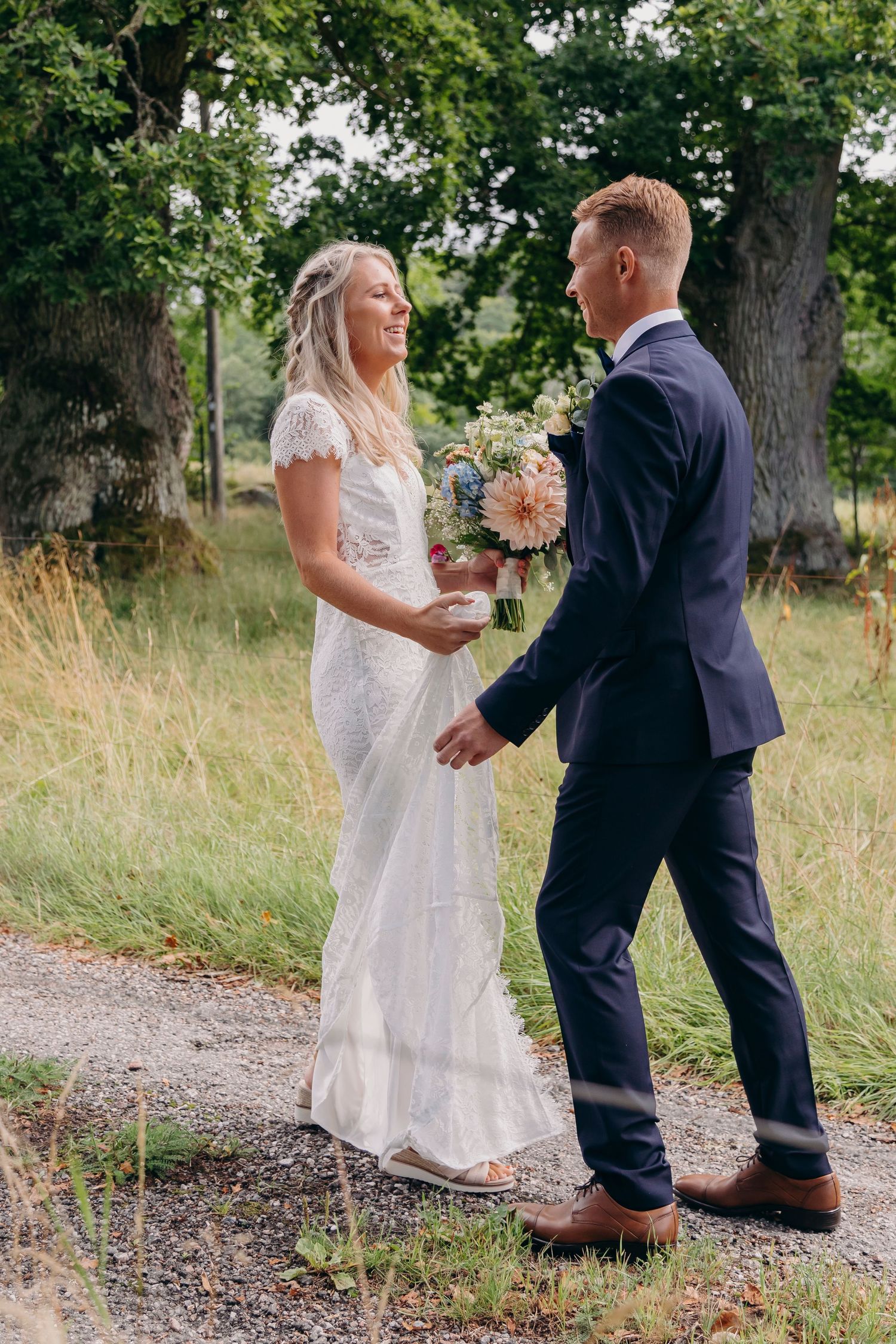 A couple in wedding attire walks together on a gravel path surrounded by tall grass and trees on a sunny day.