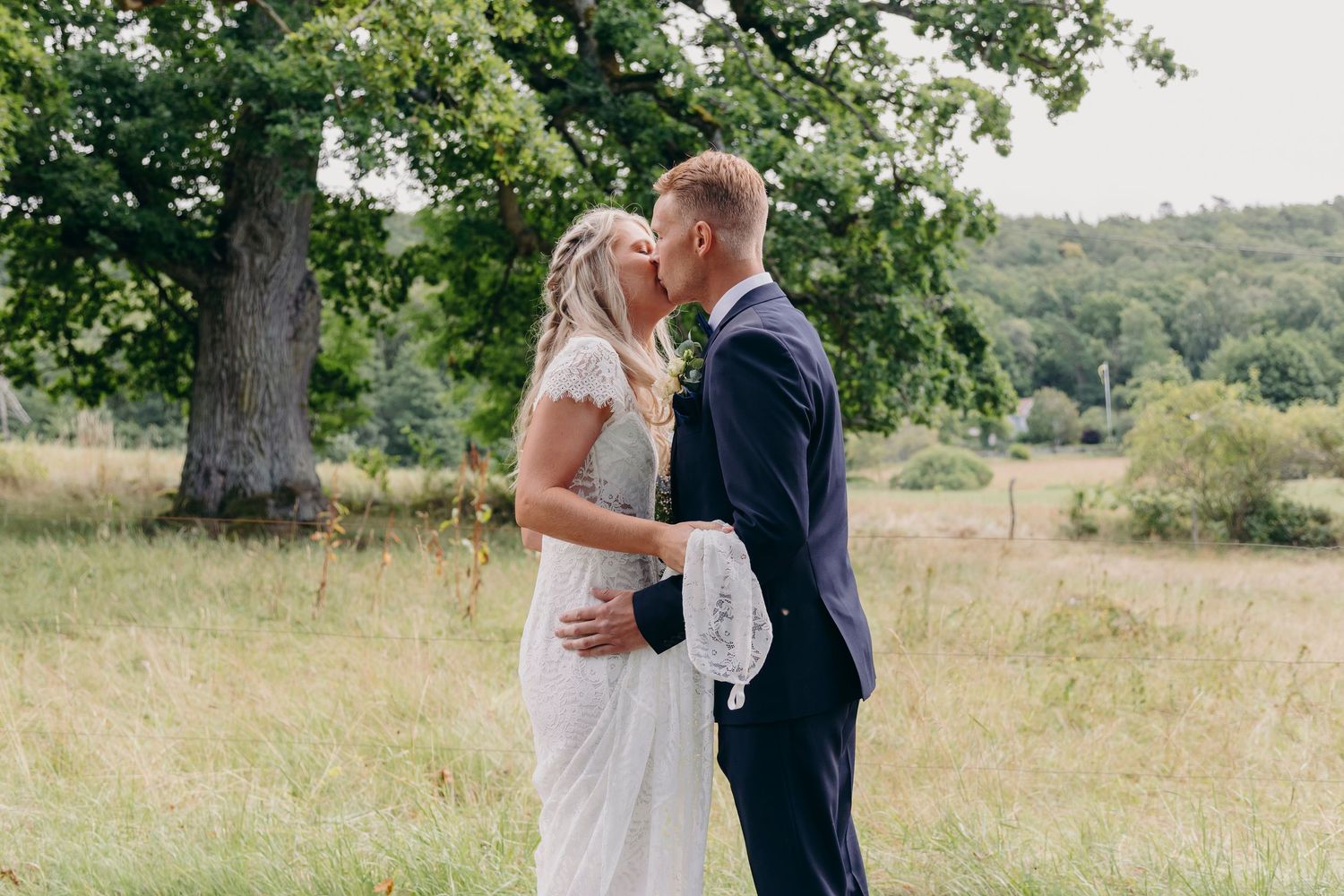 A couple shares a romantic kiss in a sunlit meadow surrounded by trees and natural greenery.