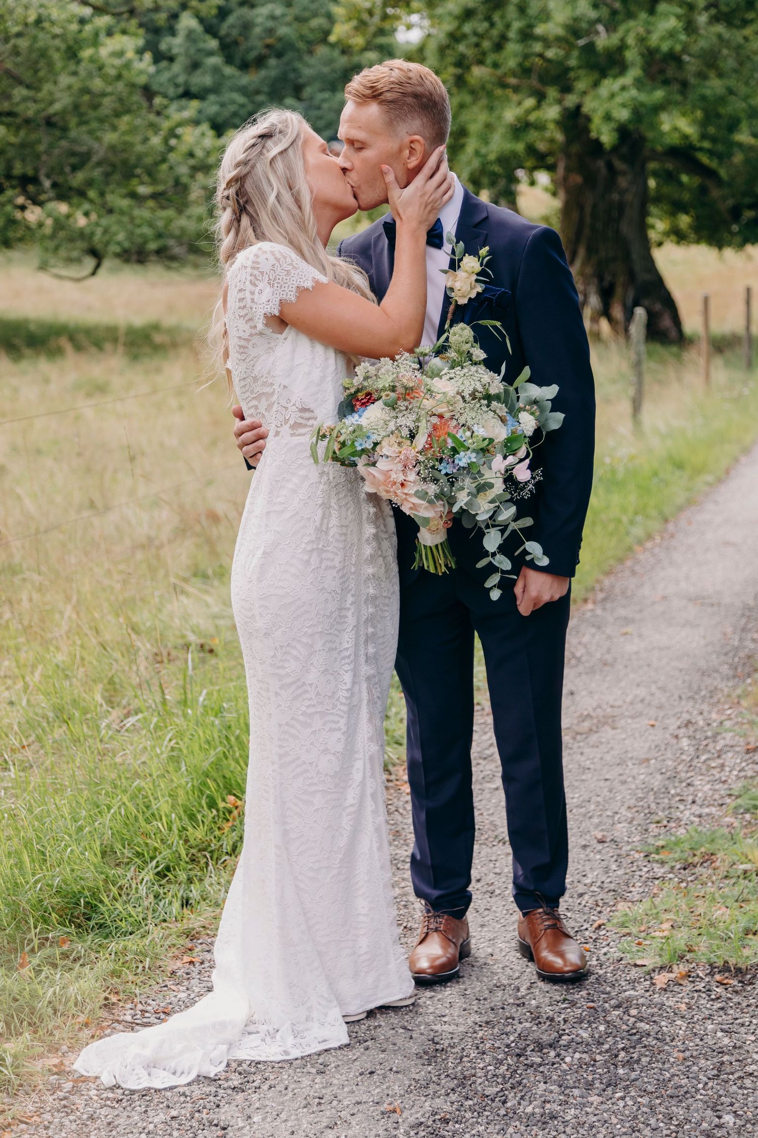 A wedding couple stands on a gravel path outdoors with the bride holding a delicate white and green bouquet.