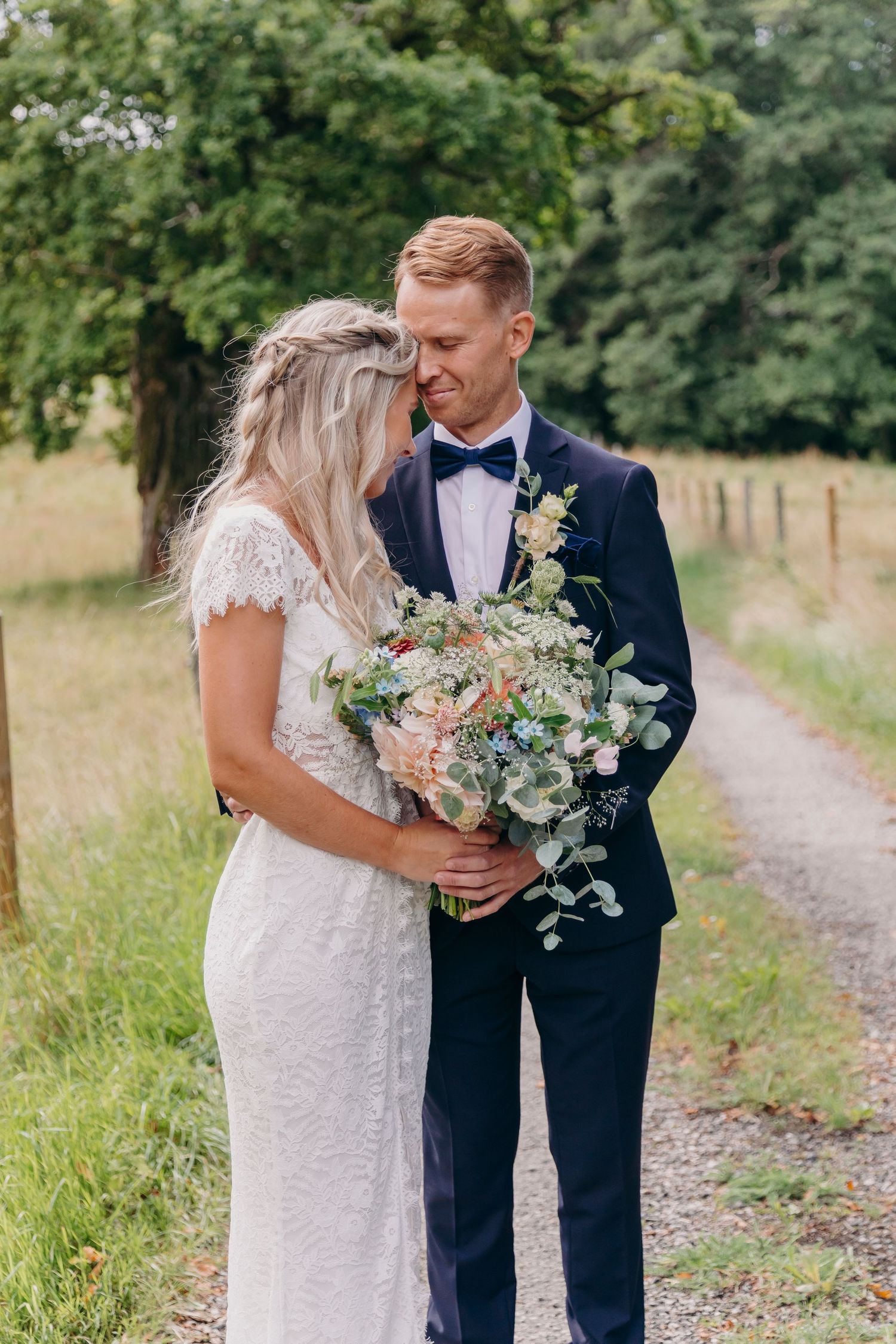 A romantic wedding couple embraces on a gravel path surrounded by lush green fields and trees on their special day.