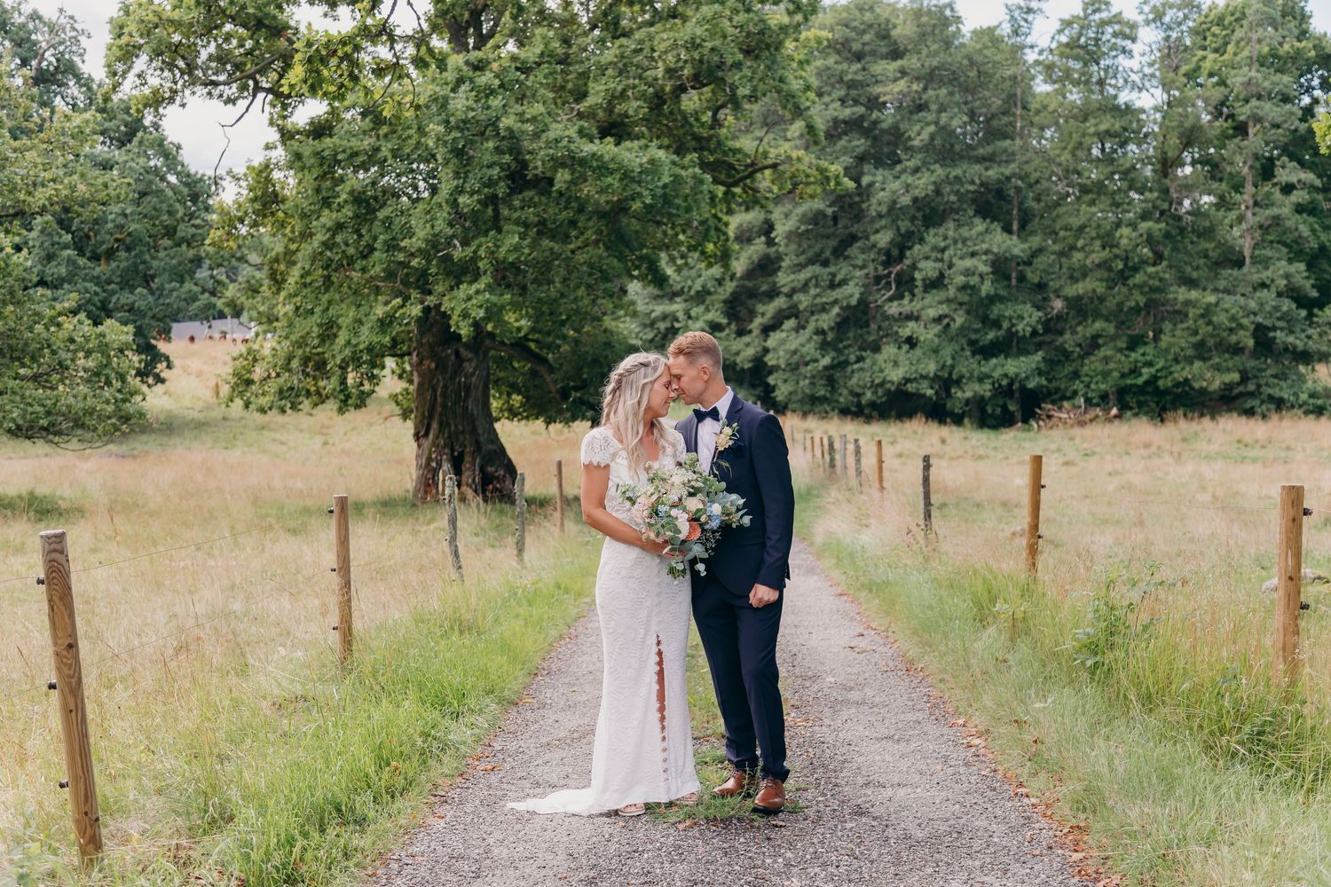 A couple walks along a scenic countryside path surrounded by lush green trees and wooden fence posts.