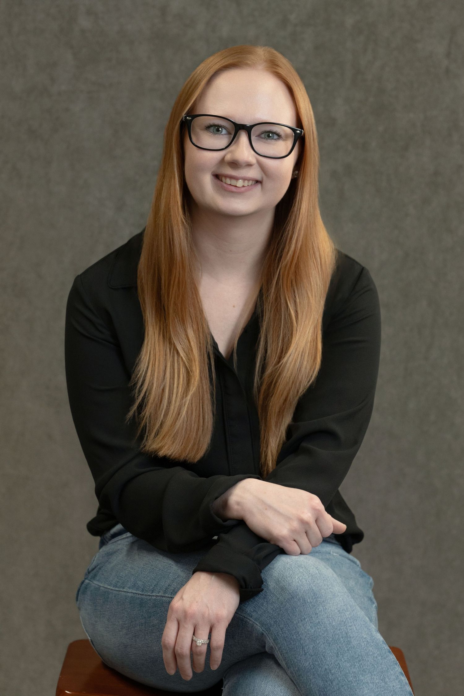 Young woman with long red hair and glasses smiling, wearing a black blouse and jeans, seated in a studio portrait.