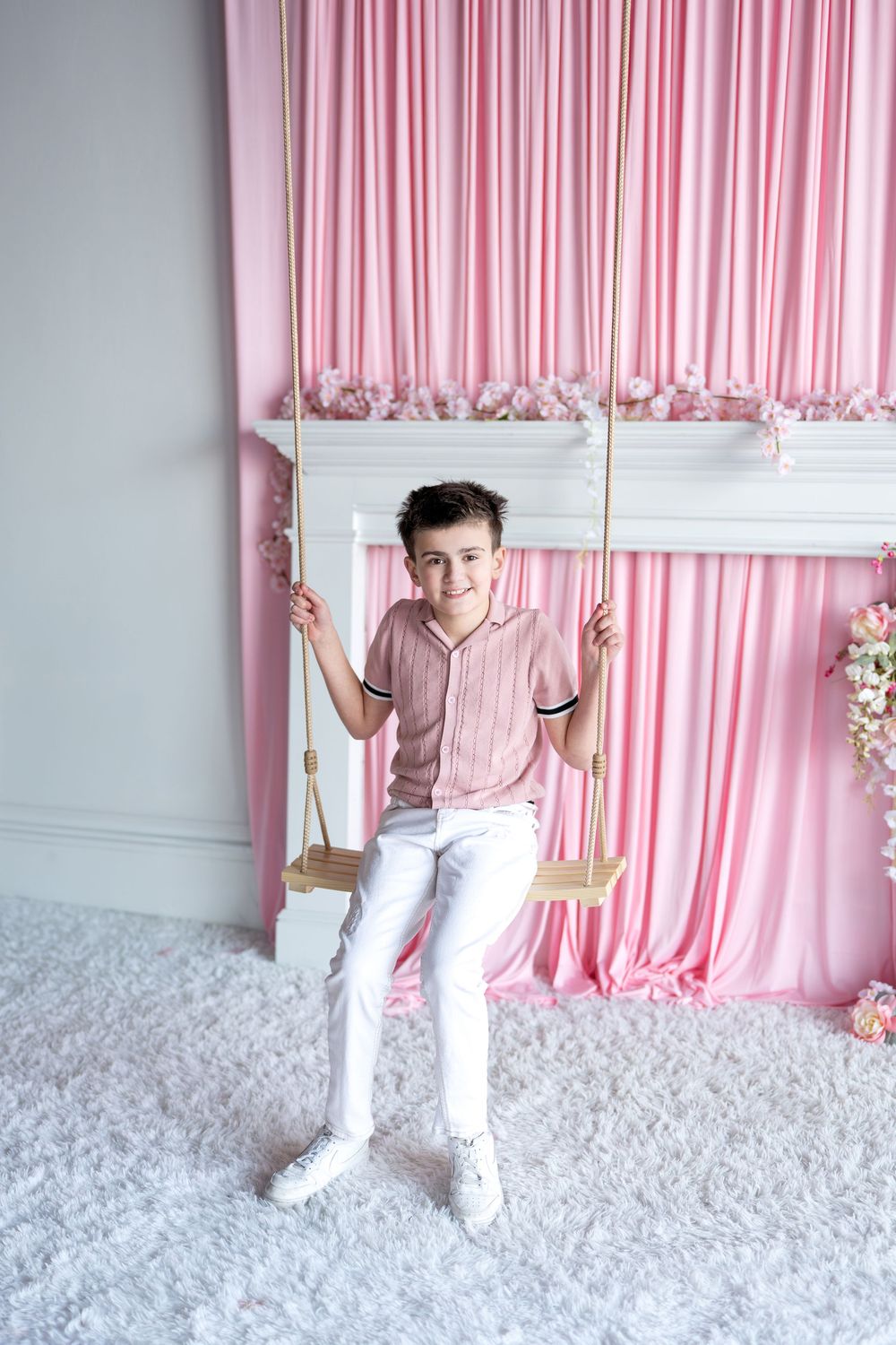 Cheerful boy in pink shirt and white pants posing on wooden swing in front of pink curtain backdrop.