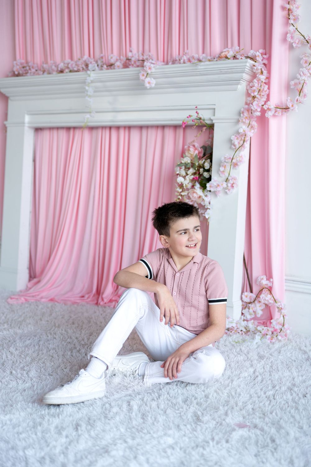 A boy in a pink shirt and white pants sits relaxed on a white fluffy rug before a pink floral backdrop.