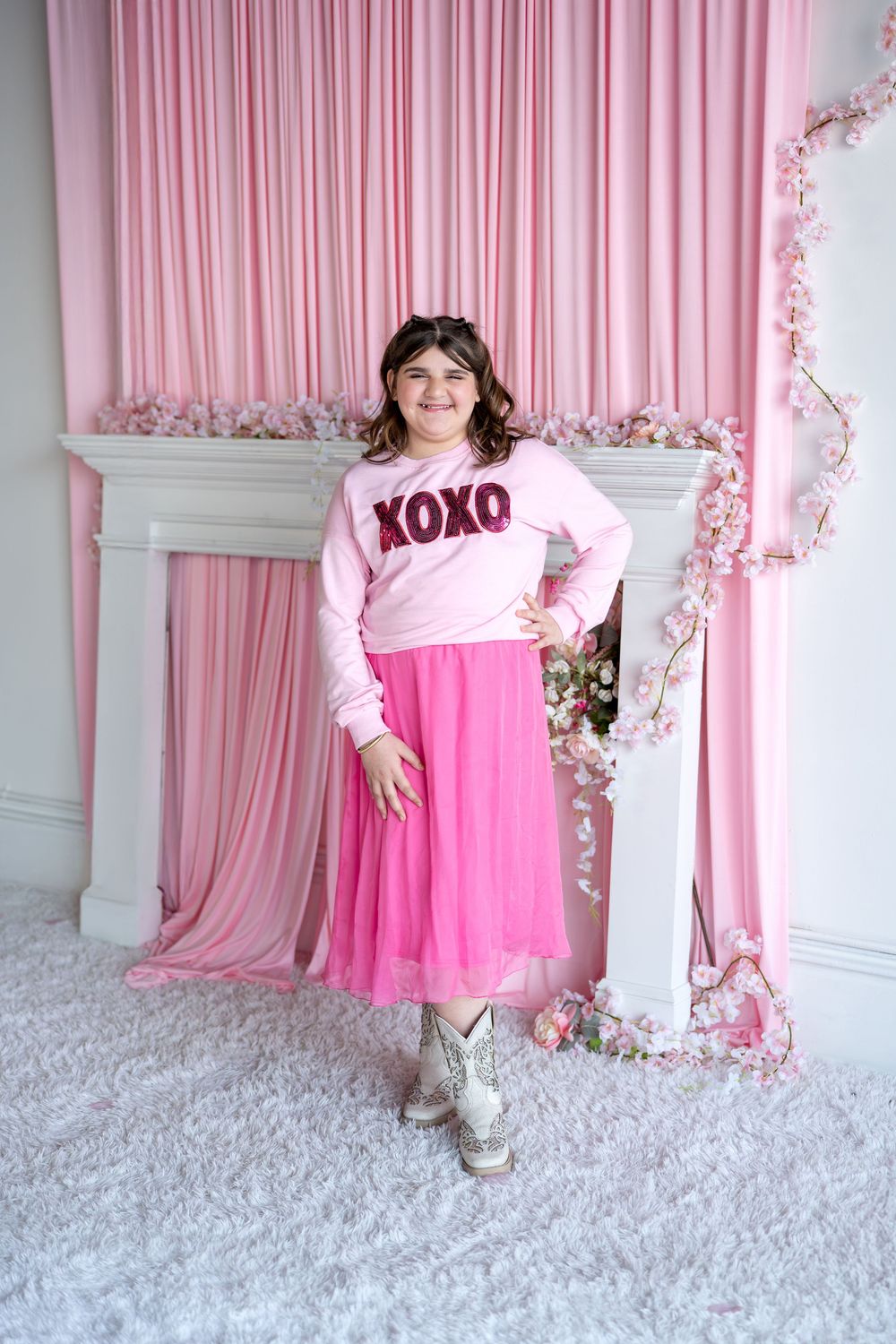 A girl in a pink XOXO sweatshirt and pink tulle skirt smiles in front of a pink curtain with flowers.
