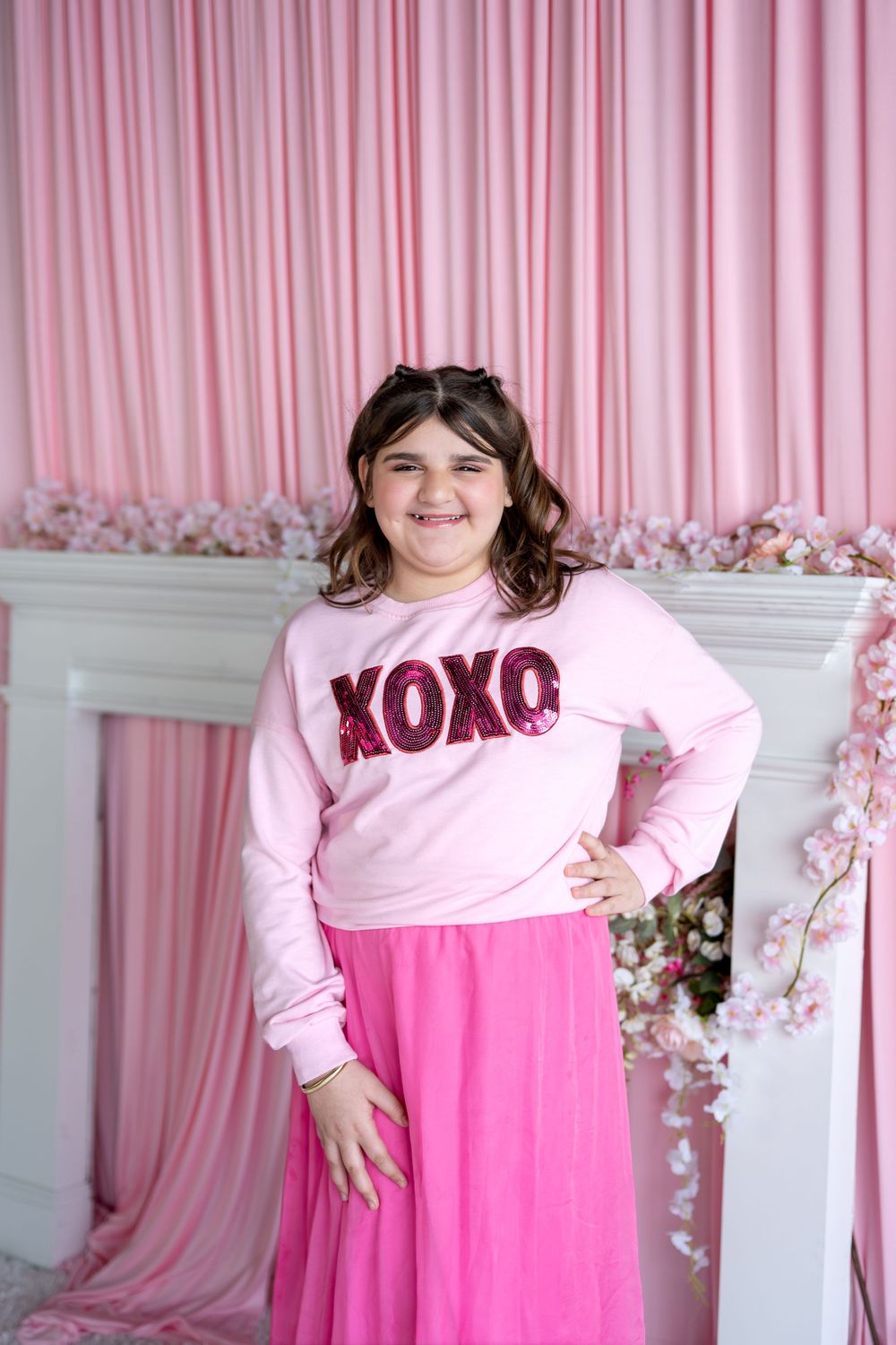 A smiling girl wearing a pink XOXO sweatshirt and bright pink tulle skirt poses in a pink floral studio.
