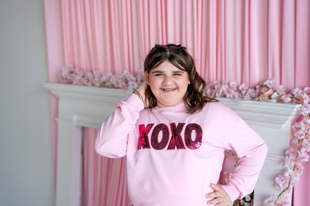 Young girl smiling in pink XOXO sequin sweatshirt against pink floral backdrop.