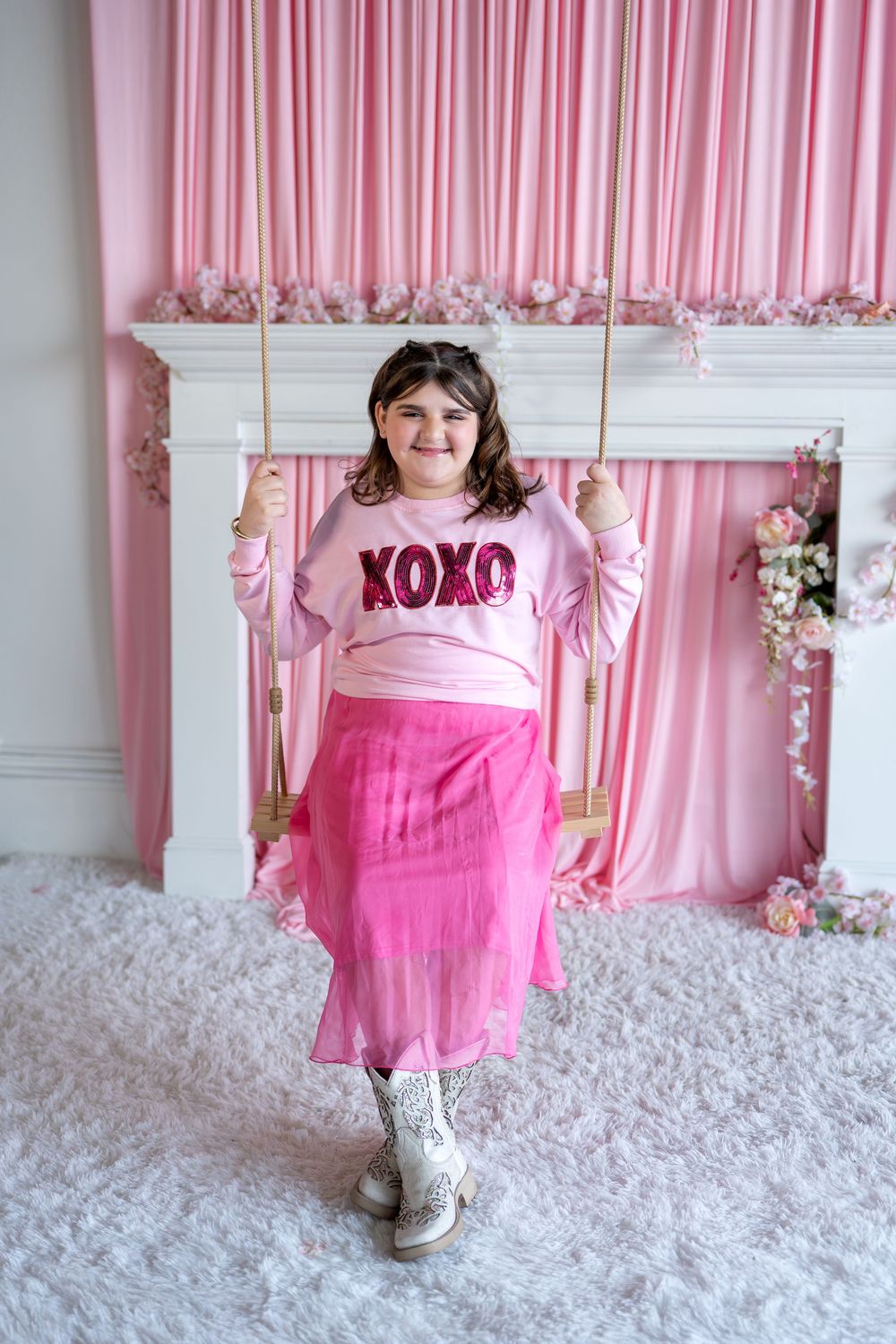 Smiling girl in pink XOXO sweatshirt and tulle skirt standing on wooden swing.
