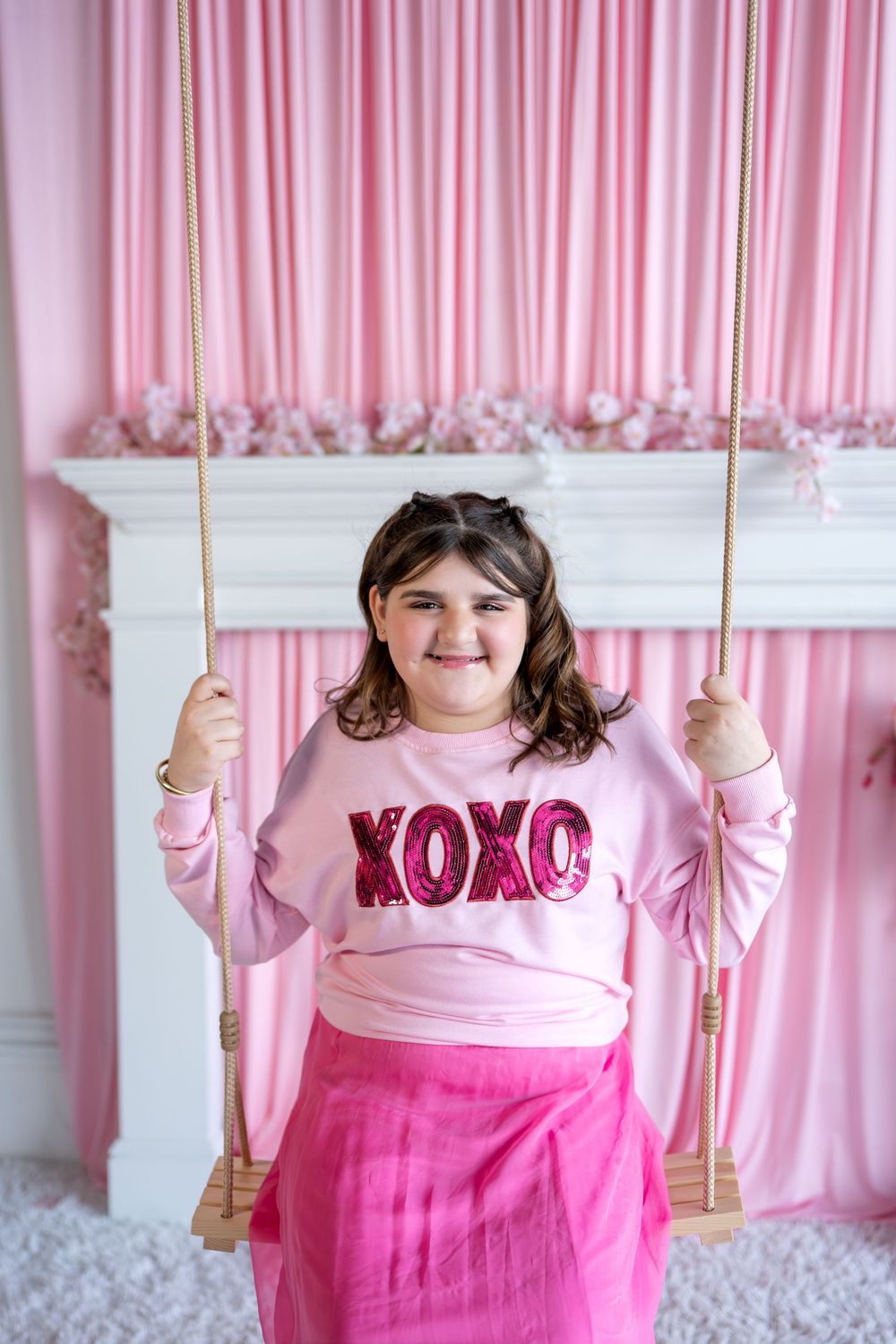 Girl in pink XOXO sweatshirt and hot pink skirt sitting on indoor wooden swing.