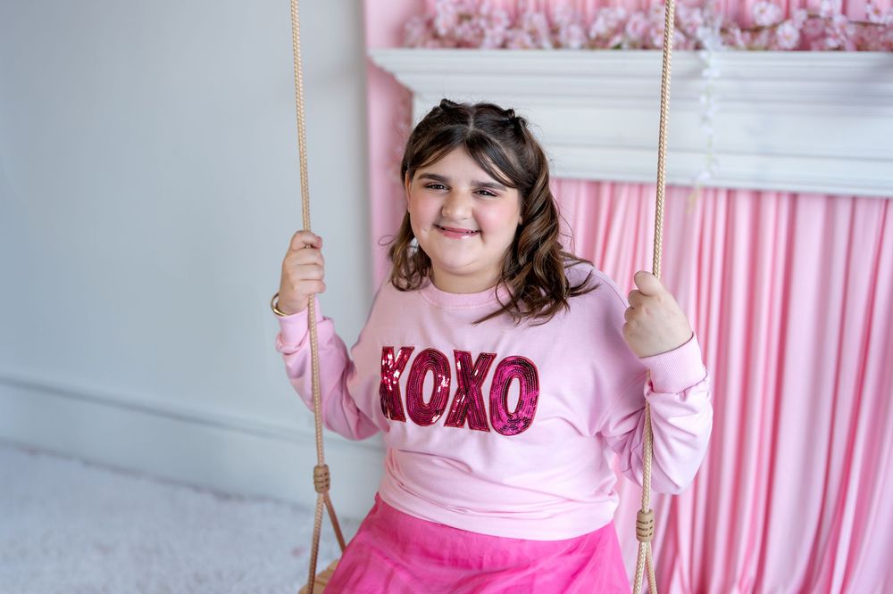 Happy girl in pink XOXO sweatshirt swinging on rope swing with pink backdrop.