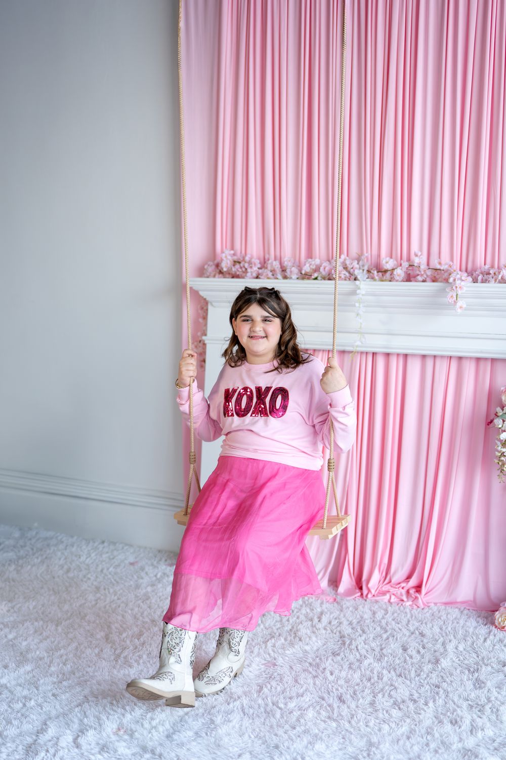 Girl in pink XOXO top and tulle skirt sitting on swing beside pink curtains.