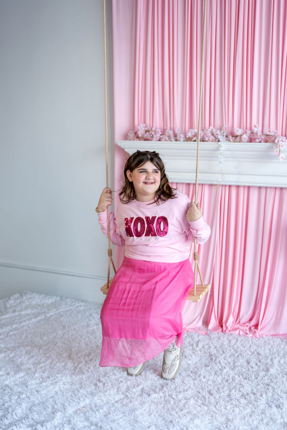 Smiling girl in pink XOXO sweatshirt and skirt sitting on wooden swing indoors.