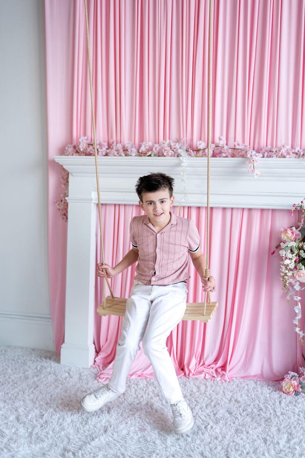 A smiling boy in a pink shirt and white pants sits on a wooden swing in a pink floral studio setting.