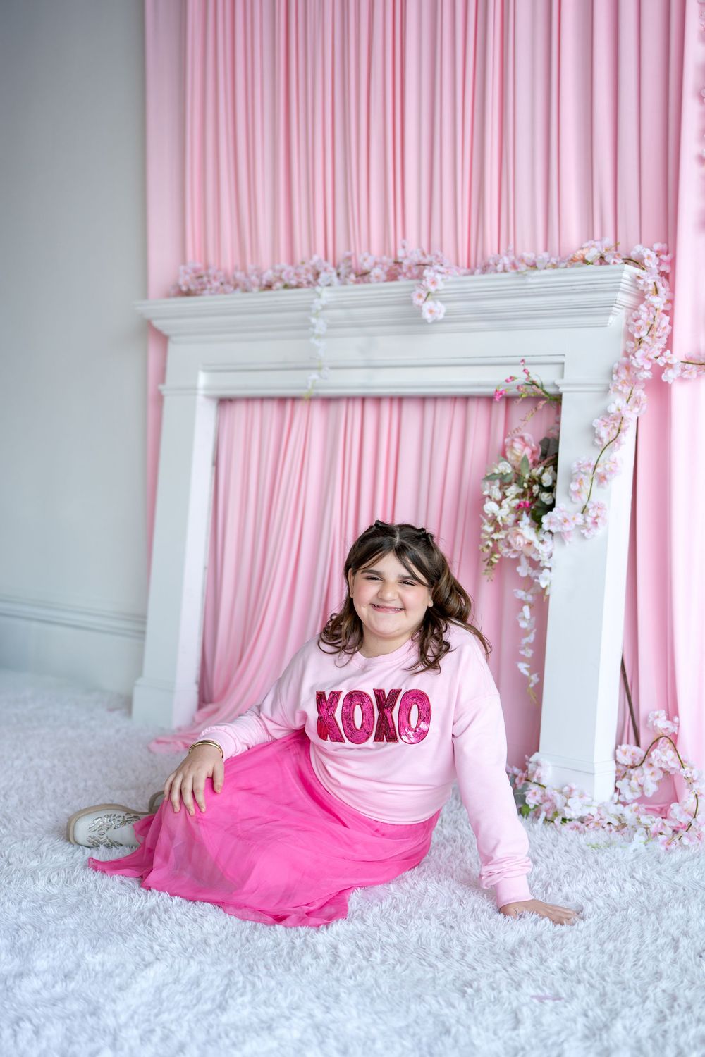 Girl in pink XOXO sweatshirt and tulle skirt sitting on white fluffy carpet.