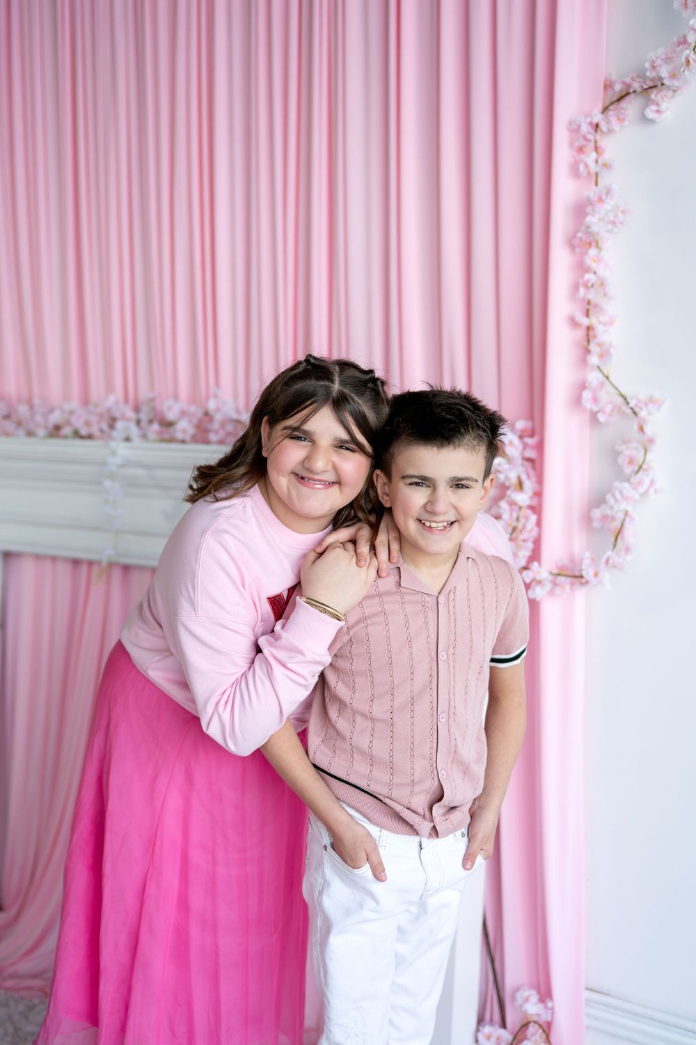 Girl in pink outfit hugging smiling boy in pink shirt and white pants indoors.