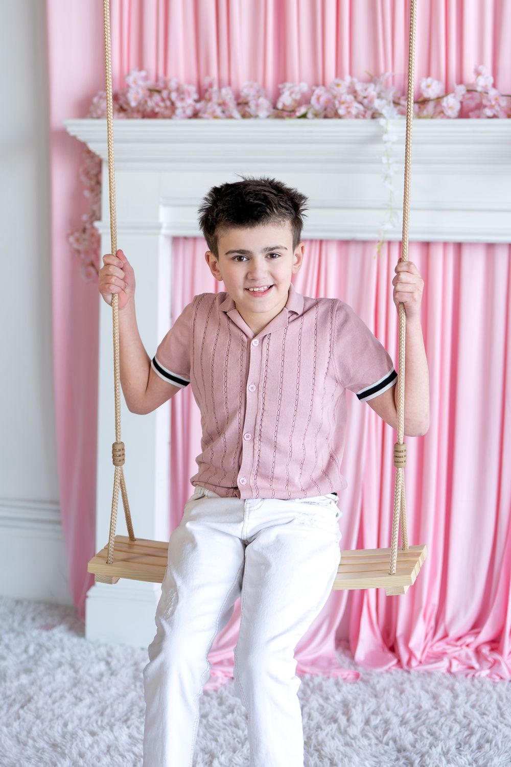 Smiling boy in pink striped shirt and white pants sitting on wooden swing in pink decorated studio.